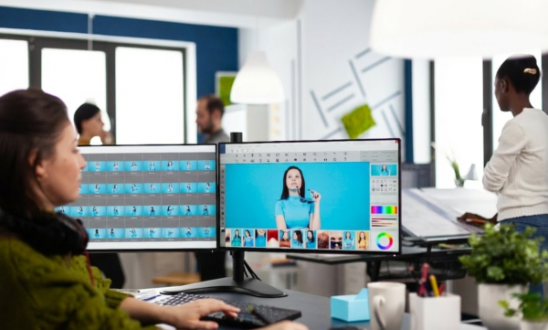 A professional content creator working at a desk with a dual-monitor setup in a modern, collaborative office. She is using graphic design software to edit a bright, colorful photo, with a grid of image variations displayed on the left screen.