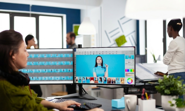 A professional content creator working at a desk with a dual-monitor setup in a modern, collaborative office. She is using graphic design software to edit a bright, colorful photo, with a grid of image variations displayed on the left screen.