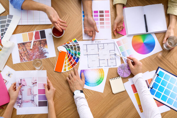 Top-down view of a creative team collaborating on a branding project. Several pairs of hands are visible around a wooden table, discussing and pointing at design materials including color swatches, mood boards, a color wheel, and layout sketches.