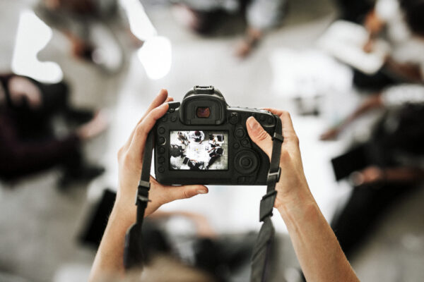 A point-of-view shot looking down at a photographer's hands holding a DSLR camera. The camera's digital display shows a top-down view of a group of people sitting in a circle on the floor, reviewing documents.