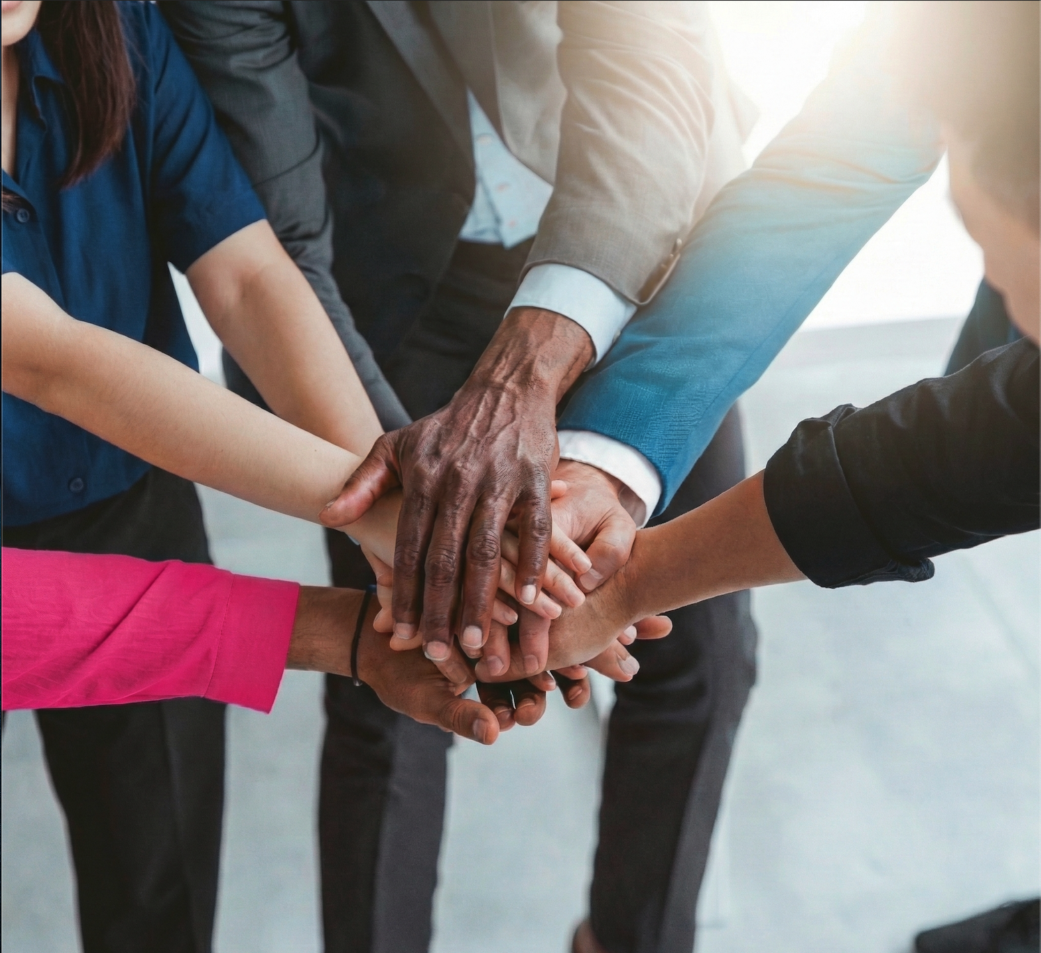 A top-down photograph of four diverse hands stacked together in a huddle, symbolizing unity and teamwork, with a bright lens flare in the upper right.